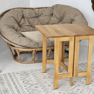 Puede incluir: Una mesa plegable de madera clara con una parte superior cuadrada y una base resistente. La mesa está colocada frente a una silla grande y redonda de mimbre con un cojín beige. También se ve una alfombra estampada y una silla blanca.