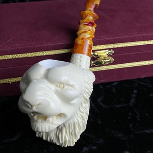 Könnte beinhalten: Eine weiße, geschnitzte Pfeife in Form eines Löwenkopfes mit einem braunen Schaft. Die Pfeife liegt auf einer burgunderfarbenen Samtbox.