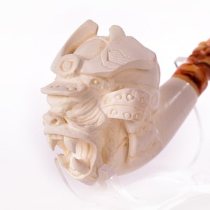 May include: A white meerschaum pipe with a detailed carving of a snarling beast's head. The pipe has a brown, marbled stem.