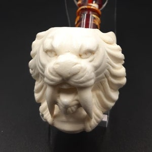 Könnte beinhalten: Eine geschnitzte Tabakpfeife in Elfenbeinfarbe mit einem detaillierten Säbelzahntigerkopf. Der Stiel der Pfeife hat eine Kombination aus Bernstein- und Rottönen. Die Pfeife wird auf einem transparenten Ständer vor dunklem Hintergrund präsentiert.