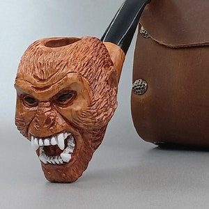 May include: A wooden pipe carved in the shape of a snarling gorilla head with a black stem. The pipe is next to a brown leather carrying case with metal accents.