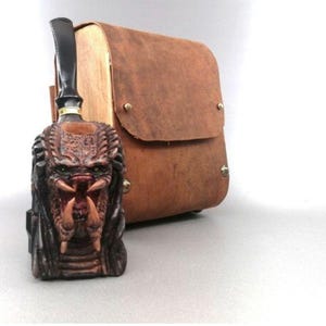 Könnte beinhalten: Eine braune Ledertasche mit einer Predator-Pfeife. Die Pfeife ist aus Holz und hat ein detailliertes Predator-Gesichtsdesign.