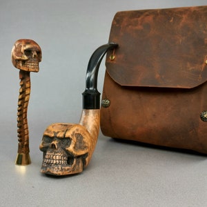 May include: A wooden smoking pipe with a skull design on the bowl and a black stem. The pipe is next to a small wooden skull with a twisted stem. A brown leather pouch with brass accents is in the background.