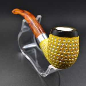 Puede incluir: Una pipa de fumar amarilla y marrón con una cazoleta texturizada. La pipa está en un soporte de plástico transparente.