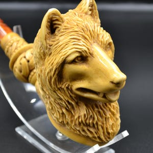 Könnte beinhalten: Eine geschnitzte Holzpfeife in Form eines Wolfskopfes. Die Pfeife ist hellbraun und hat eine detaillierte Schnitzerei des Wolfsfells und der Gesichtszüge. Die Pfeife steht auf einem durchsichtigen Acrylständer.