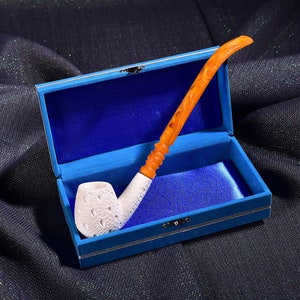 Puede incluir: Una pipa blanca con un diseño tallado y una boquilla naranja está en una caja de terciopelo azul con ribete plateado.
