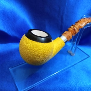 Puede incluir: Una pipa amarilla y marrón con una cazoleta texturizada y un borde negro. La pipa está en un soporte transparente.