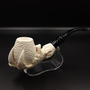May include: A white ceramic pipe shaped like a dragon's claw with a black mouthpiece. The pipe is on a clear acrylic stand.