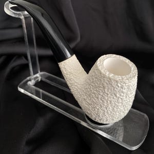 Könnte beinhalten: Eine weiße Keramikpfeife mit einem schwarzen Mundstück, auf einem transparenten Acrylständer ausgestellt. Die Pfeife hat eine strukturierte Oberfläche.