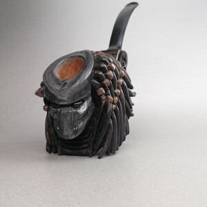 Könnte beinhalten: Eine schwarze und silberne Predator-Pfeife mit einem Holzmundstück und einem schwarzen Schaft. Die Pfeife ist so geformt, dass sie wie der Kopf des Predators mit Dreadlocks und einer Maske aussieht.