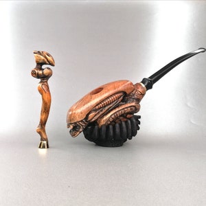 May include: A wooden pipe carved in the shape of an alien head with a black stem. The pipe is on a black stand. A small wooden figurine is also in the image.