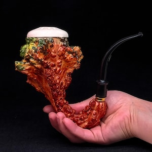 Puede incluir: Una pipa de tabaco artesanal con un diseño texturizado único. La pipa presenta un cuenco de color marrón rojizo, de aspecto orgánico, con una tapa blanca y una boquilla negra curvada. La pipa se sostiene en una mano sobre un fondo oscuro.