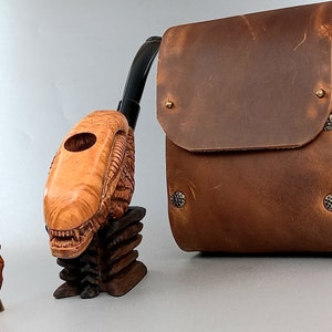 May include: A wooden pipe shaped like an alien head with a brown leather carrying case. The pipe has a carved design and a hole for the tobacco. The case has a flap and a snap closure.