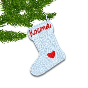 May include: A light blue Christmas stocking with the name "Коста" embroidered in red. The stocking features a red heart and a decorative pattern. A green pine branch is in the background.