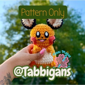 Könnte beinhalten: Ein gehäkeltes Plüschtier mit gelbem Körper, weißem Bauch und großen Ohren mit schwarzen und weißen Details. Das Gesicht hat schwarze Augen, rosa Wangen und einen kleinen Mund. Der Text lautet Pattern Only und @Tabbigans.