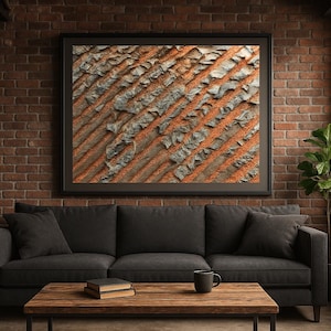 May include: A framed abstract art print with a textured, weathered appearance. The artwork features diagonal lines of orange and gray, creating a rustic aesthetic. The print hangs above a dark gray sofa in a living room setting.