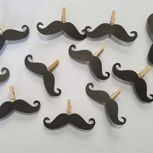 May include: A set of twelve black paper mustache cutouts with wooden clothespins attached. The mustaches are all the same size and shape, and they are arranged in a random pattern on a white background.