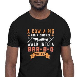 Op de afbeelding: Een zwart T-shirt met een wit grafisch ontwerp met de tekst "A cow, a pig and a chicken walk into a bar-b-q the end".