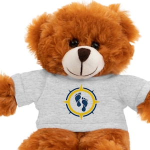 Könnte beinhalten: Ein brauner Teddybär mit einem hellgrauen T-Shirt. Das T-Shirt zeigt ein Kompassdesign mit blauen Fußabdrücken in einem gelben Kreis. Der Bär hat eine hellbraune Schnauze und dunkle Augen, die Arme sind ausgestreckt.