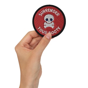 May include: A red and black embroidered patch with a white skull and crossbones and the text "SURRENDER YOUR BOOTY".