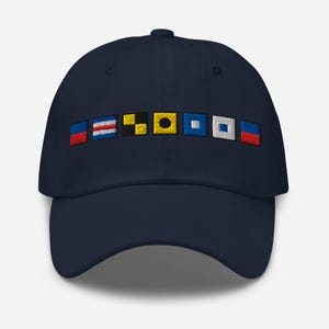 May include: Navy blue baseball cap with embroidered nautical signal flags in various colors. The flags are arranged horizontally across the front of the cap. The cap has a curved brim and a button on top.