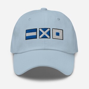 May include: Light blue baseball cap with embroidered nautical signal flags in blue and white. The cap features a curved brim and an adjustable strap at the back. The flags are a rectangle with a horizontal stripe, an X, and a square.