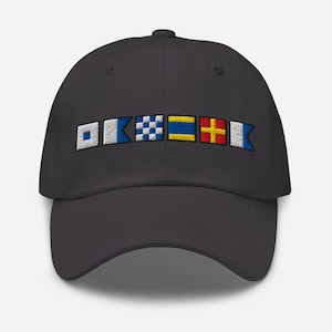 Può includere: Un cappellino da baseball grigio scuro con bandiere nautiche ricamate in bianco, blu, giallo e rosso. Le bandiere sono disposte orizzontalmente sulla parte anteriore del cappellino. Il cappellino ha una visiera curva e un bottone sulla parte superiore.