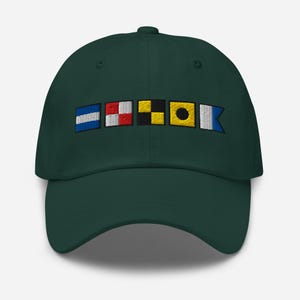 Puede incluir: Gorra de béisbol verde oscuro con banderas de señales náuticas bordadas en azul, rojo, negro, amarillo y blanco. La gorra tiene una visera curva y un cierre de correa de tela.