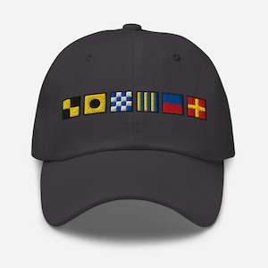 May include: A dark gray baseball cap featuring embroidered nautical signal flags in various colors. The flags include black and yellow, yellow and black, blue and white checkered, yellow stripes, blue and red, and red and yellow cross designs.