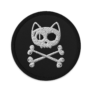 以下が含まれることがあります： 猫の頭蓋骨と交差した骨が刺繍された黒と白の刺繍パッチ。パッチは円形で、黒い縁取りがあります。