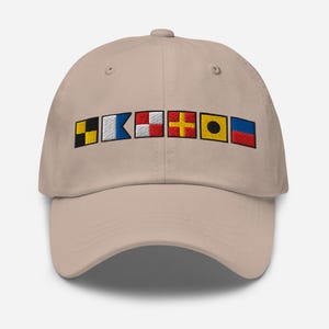 Puede incluir: Gorra de béisbol beige con banderas de señales náuticas bordadas en negro, amarillo, rojo, blanco y azul. La gorra tiene una visera curva y un cierre de correa de tela en la parte trasera.