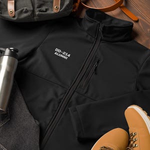 May include: Black softshell jacket with a zipper closure and a zippered pocket on the left side. The jacket has the text "DD-214 ALUMNI" embroidered on the left chest.