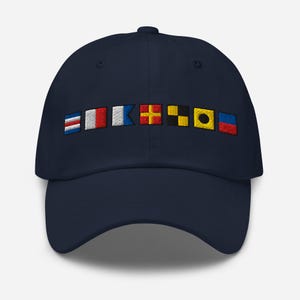 Puede incluir: Gorra de béisbol azul marino con banderas de señales náuticas bordadas en varios colores. Las banderas están dispuestas horizontalmente en la parte delantera de la gorra. La gorra tiene una visera curva y un botón en la parte superior.