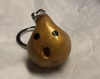 Könnte beinhalten: Ein goldfarbener, birnenförmiger Schlüsselanhänger mit einem schwarz bemalten Gesicht. Der Schlüsselanhänger hat zwei schwarze Augen und einen weit geöffneten Mund.