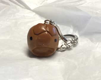 Könnte beinhalten: Ein brauner, runder Schlüsselanhänger mit einem Gesicht und einer silbernen Kette. Der Schlüsselanhänger hat zwei schwarze Augen und einen kleinen Mund.