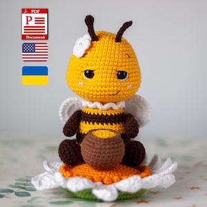 Op de afbeelding: Een gehaakt bijenspeeltje met een geel en bruin gestreept lichaam, zittend op een witte en oranje bloem. De bij heeft een witte bloem als accessoire op het hoofd, twee voelsprieten en houdt een kleine bruine honingpot vast. Het speelgoed heeft geborduurde ogen.