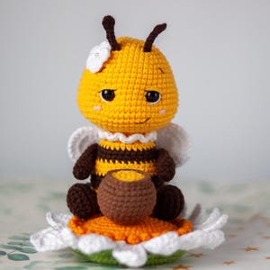May include: A handmade crochet bee figurine with a yellow body, brown stripes, and white wings. The bee is sitting on a flower with white petals, an orange center, and a green base. It holds a small brown pot.