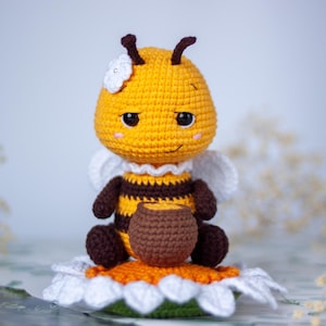 Pode incluir: Uma boneca de abelha em crochê com cabeça amarela, corpo listrado de amarelo e marrom e membros marrom-escuros. A abelha possui uma flor branca na cabeça, asas brancas e segura um pequeno pote de mel marrom sentada sobre uma grande margarida.