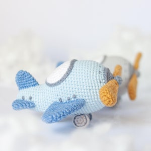 Op de afbeelding: Twee gehaakte vliegtuig speelgoedjes, een blauwe en een grijze, met oranje propellers. Het blauwe vliegtuig is scherp en het grijze vliegtuig is wazig op de achtergrond.