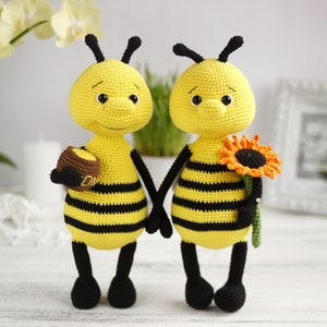 以下が含まれることがあります： 2つのクロシェ編みミツバチのぬいぐるみ。1つは小さなハチミツの壺を持っており、もう1つは黄色のひまわりを持っています。ミツバチは黄色と黒で、黒い目と笑顔をしています。
