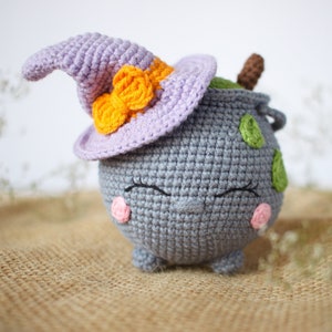 May include: A gray crocheted cauldron with a purple witch hat and orange bow. The cauldron has green and white accents and a cute face with pink cheeks.