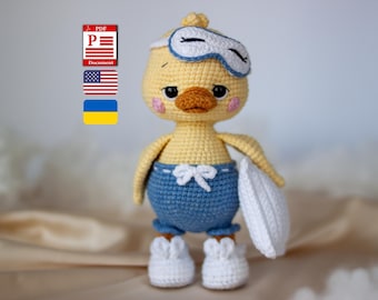 May include: A yellow crocheted duck doll wearing blue shorts with a white drawstring, white shoes, and a white sleep mask over its eyes. The duck holds a small white pillow in one hand. The figure has pink cheeks and a brown beak.