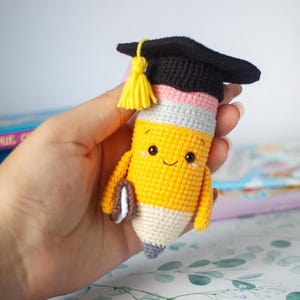 May include: A handmade, crocheted pencil-shaped plush toy with a smiling face, wearing a black graduation cap with a yellow tassel. The pencil has a yellow body, a pink and grey section, and a white tip. It holds a small book.