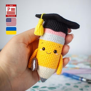 May include: A handmade crocheted pencil doll wearing a graduation cap. The pencil is yellow, pink, grey, and black, with a smiling face and small arms. The graduation cap is black with a yellow tassel. The doll is held in a hand.