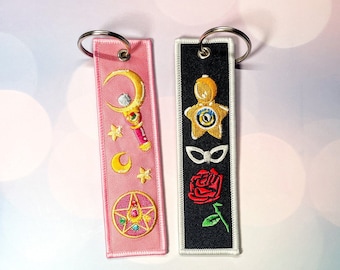 Peut inclure: Deux porte-clés rose et noir avec des images brodées. Le porte-clés rose a un croissant de lune, des étoiles, une baguette et une étoile avec un cristal au centre. Le porte-clés noir a une étoile avec un œil au centre, un masque, une rose rouge et une feuille verte.