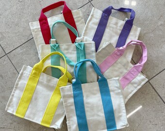 Könnte beinhalten: Ein Set aus sechs kleinen Tragetaschen mit bunten, gestreiften Griffen. Die Taschen sind aus weißem Canvas gefertigt und haben eine Vielzahl von Farben, darunter Rot, Lila, Rosa, Gelb, Blau und Grün.
