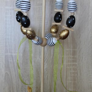 May include: An Easter egg wreath made of wood, featuring eggs painted in black, white, and gold. Some eggs have polka dots, others stripes. The wreath is supported by a wooden stand and decorated with green ribbons and a small bunny figurine.