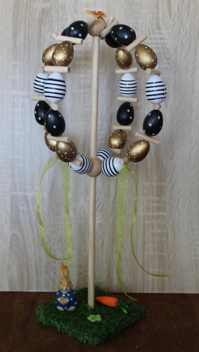 May include: An Easter egg wreath made of wood, featuring eggs painted in black, white, and gold. Some eggs have polka dots, others stripes. The wreath is supported by a wooden stand and decorated with green ribbons and a small bunny figurine.