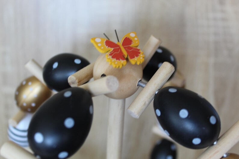 May include: Wooden Easter egg tree featuring black eggs with white polka dots, and a gold egg with white polka dots. A yellow and red butterfly decorates the top. The tree is made of wood with a light brown background.