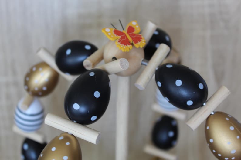 May include: A decorative Easter egg display made of wood, featuring black and gold eggs with white polka dots. A small butterfly with red and yellow wings is at the top. The eggs are arranged on wooden dowels.